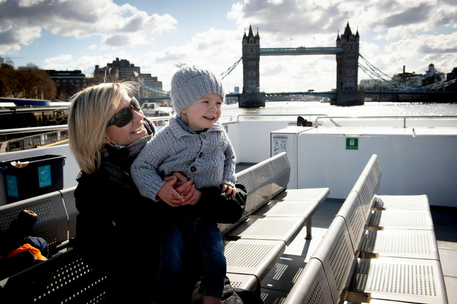 Woman holding a smiling child on a boat with the Tower Bridge in the background on a partly cloudy day.