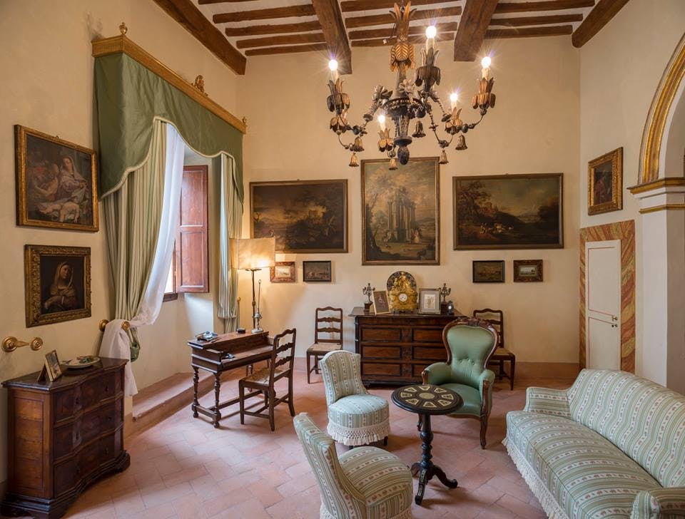 Un salotto vintage con mobili antichi, un lampadario, quadri alle pareti e una finestra con tende verdi e bianche.