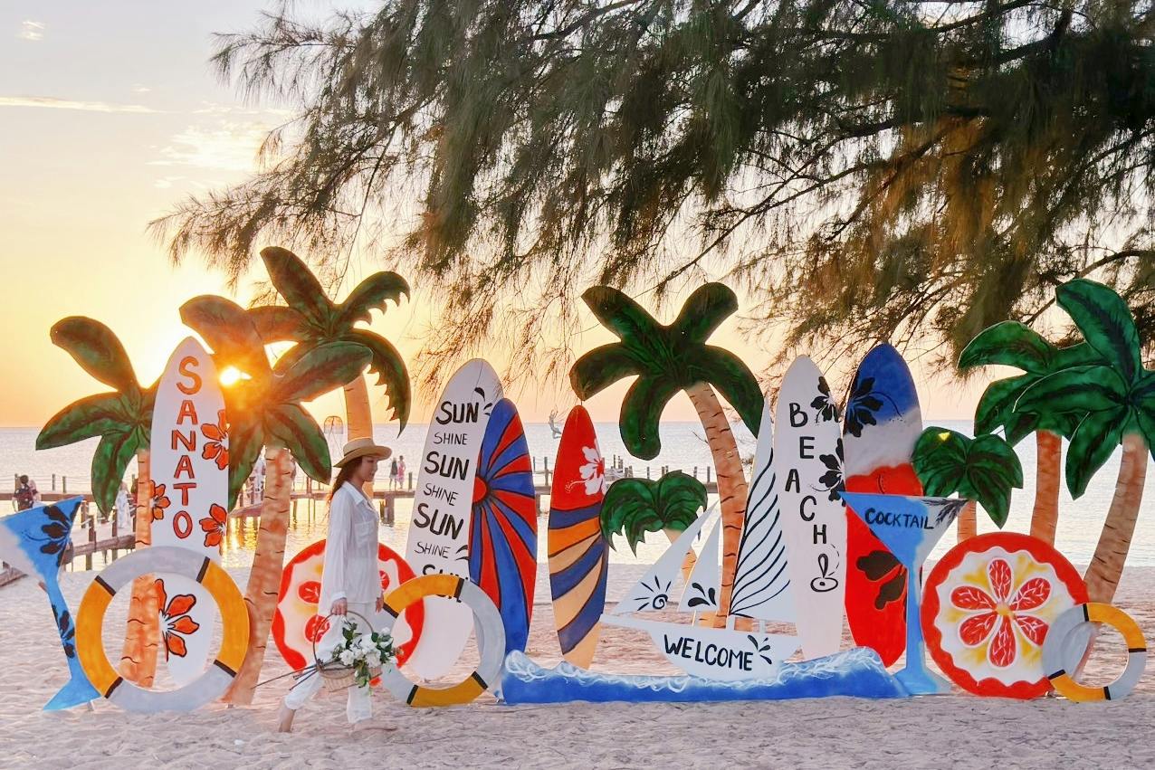 Scena plażowa z deskami surfingowymi, palmami i przedmiotami dekoracyjnymi ułożonymi na piasku o zachodzie słońca, z osobą przechodzącą obok.