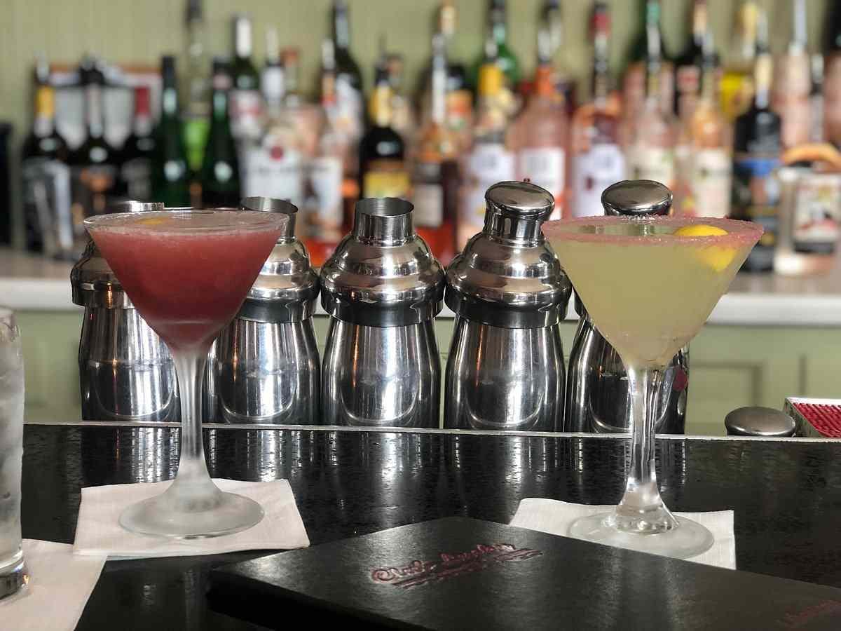 Two cocktails on a bar counter, one red and one yellow, with shakers and various liquor bottles in the background.