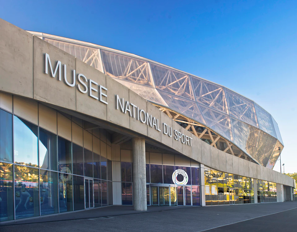 Modern building with "MUSEE NATIONAL DU SPORT" sign, featuring glass panels and a geometric roof structure under clear blue sky.