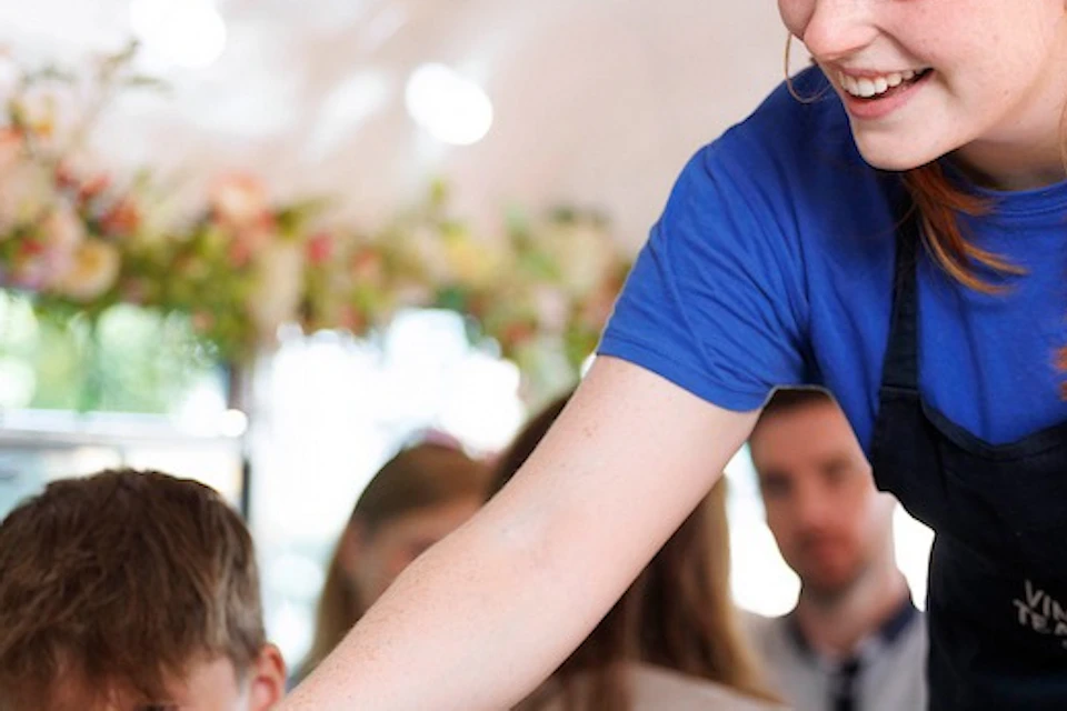 A smiling woman in a blue shirt serves food from a basket to seated people on a bus decorated with flowers.