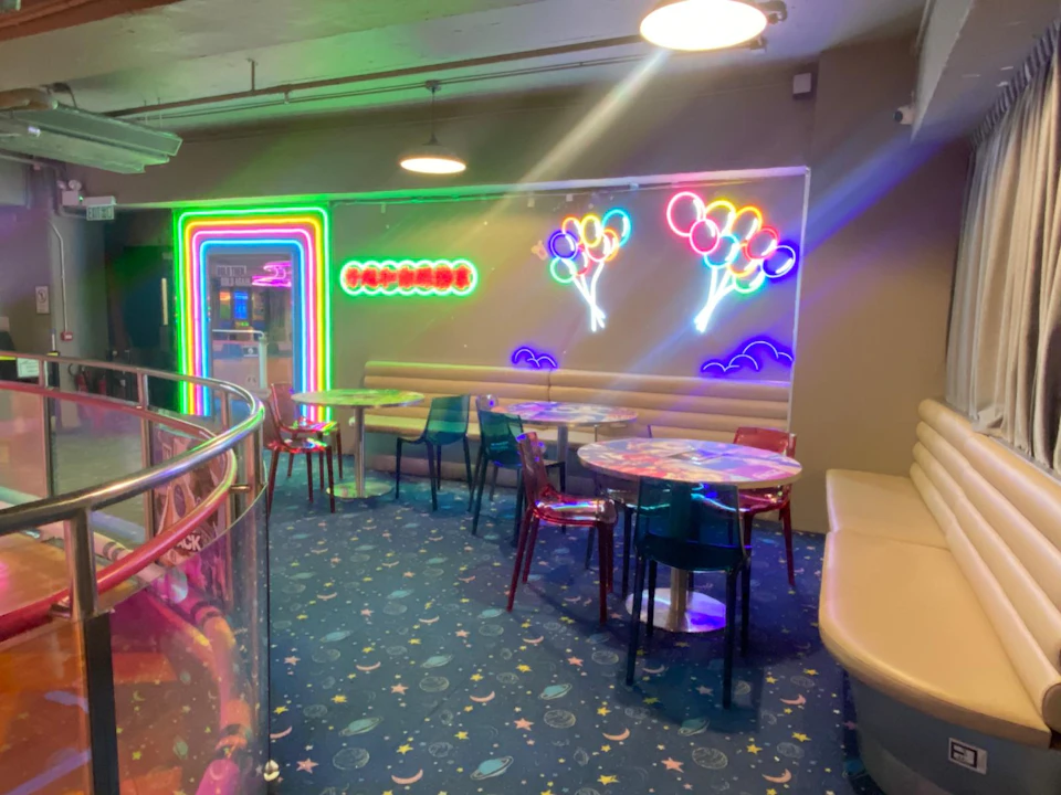 A colorful indoor seating area with neon balloon lights, a rainbow arch, tables, chairs, and a space-themed carpet.