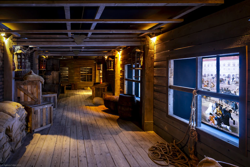 Interior of a wooden ship or themed room with barrels, crates, ropes, and lanterns under warm lighting.