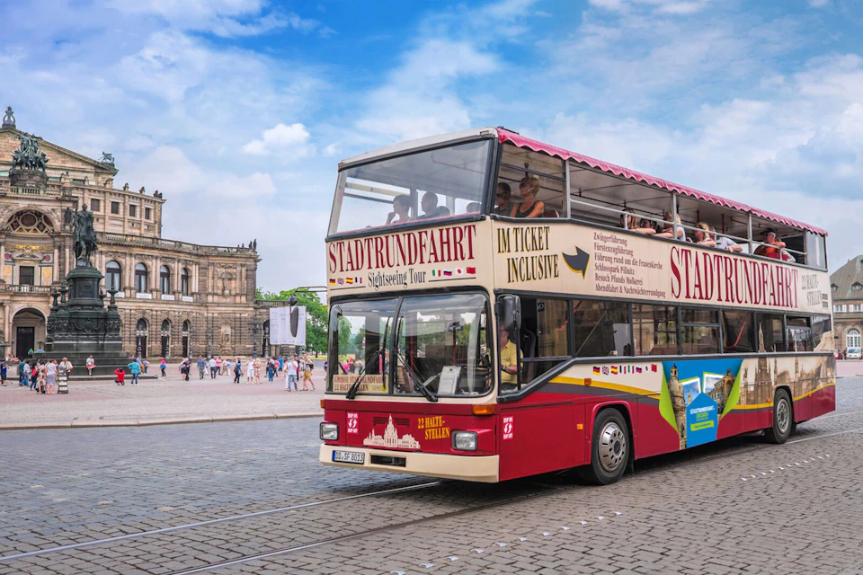 Double-decker sightseeing bus with German text on its side in a historic city square with people and an ornate building in the background.