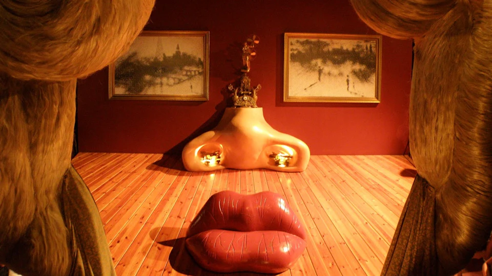 Art installation featuring a lip-shaped couch, a nose-shaped sculpture, and two paintings on a wooden floor with a red wall.