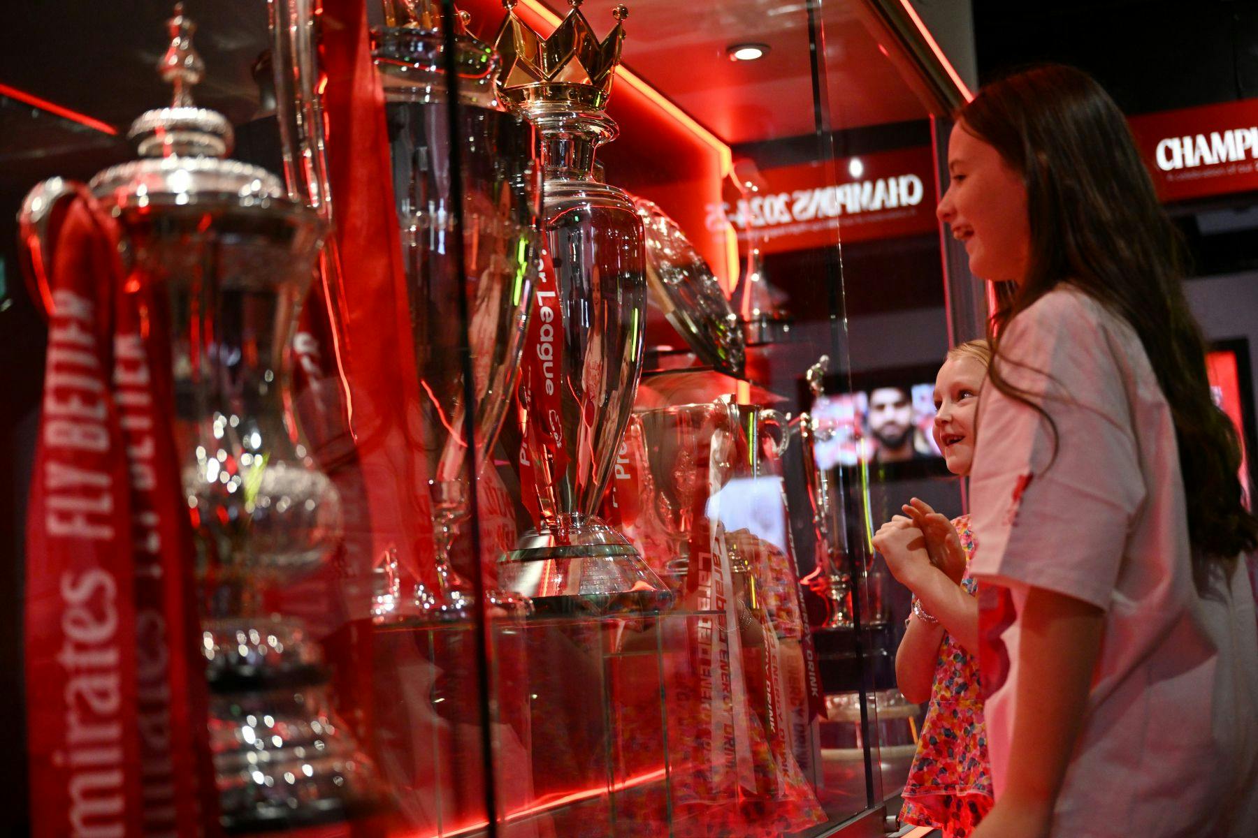 두 어린 소녀는 빨간색 조명이 있는 유리 케이스에 전시된 커다란 은색 트로피에 감탄합니다.