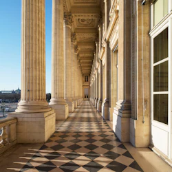 Utsmyckad korridor med höga pelare, detaljerade tak och ett svartvitt rutigt golv, sett i dagsljus.