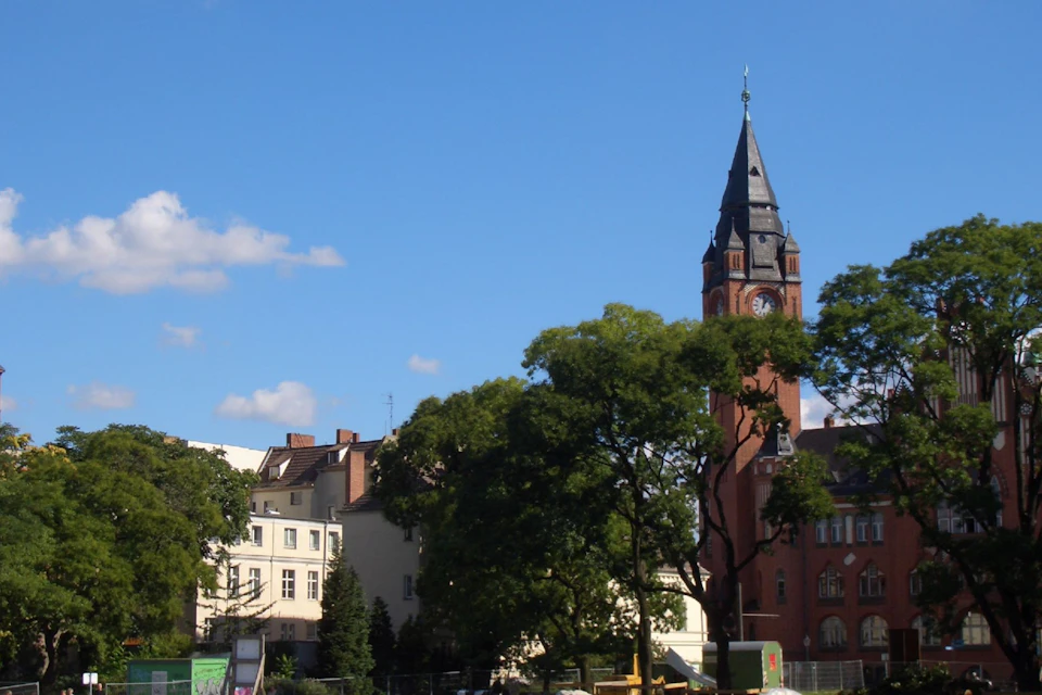 A tall clock tower with a steep roof stands near trees and buildings under a clear blue sky.