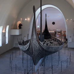 An ancient wooden Viking ship displayed in a museum, surrounded by ropes and barriers with an arched ceiling above.