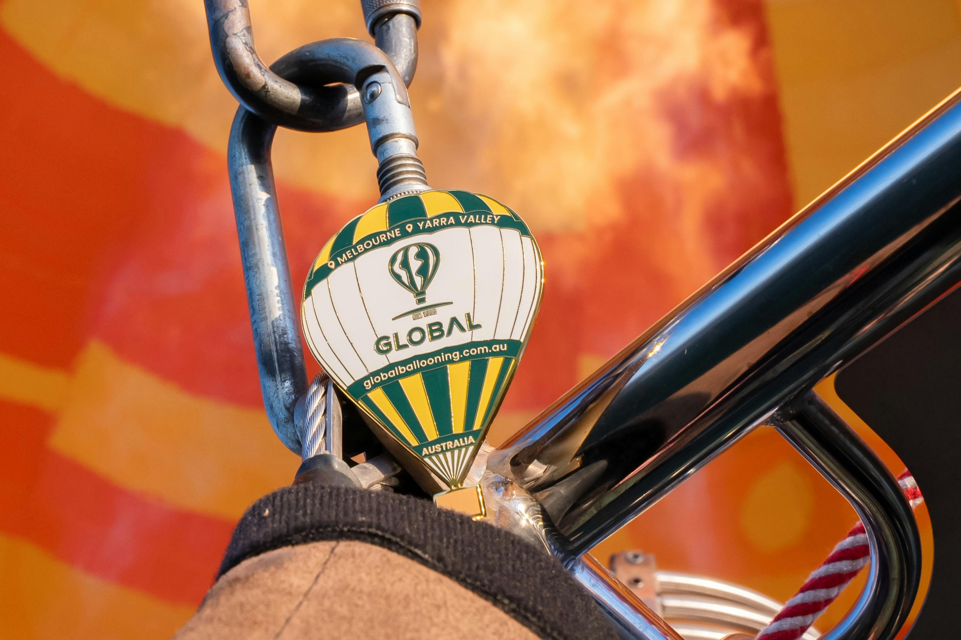 Close-up of a keychain shaped like a hot air balloon, hanging from a metal hook with a blurred colorful background.