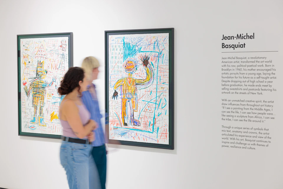 Two people observe framed artwork by Jean-Michel Basquiat in an exhibition space with explanatory text on a wall.