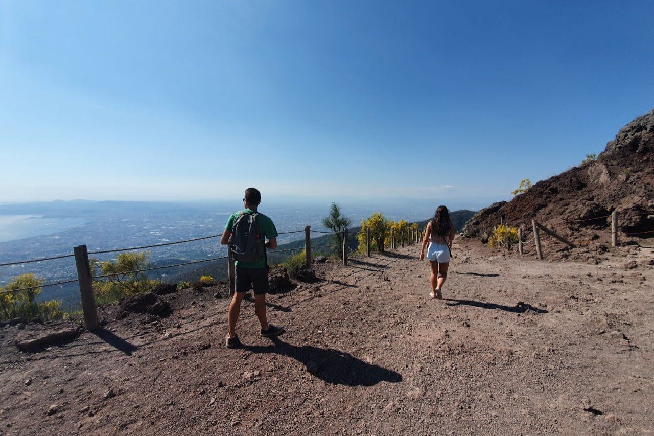 Deux personnes se promènent sur un chemin de terre avec une ville et un ciel bleu en arrière-plan, bordé par une clôture en bois et une végétation clairsemée.