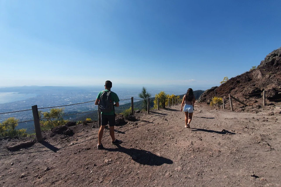 Two people hiking on a dirt trail with a city and blue sky in the background, bordered by a wooden fence and sparse vegetation.