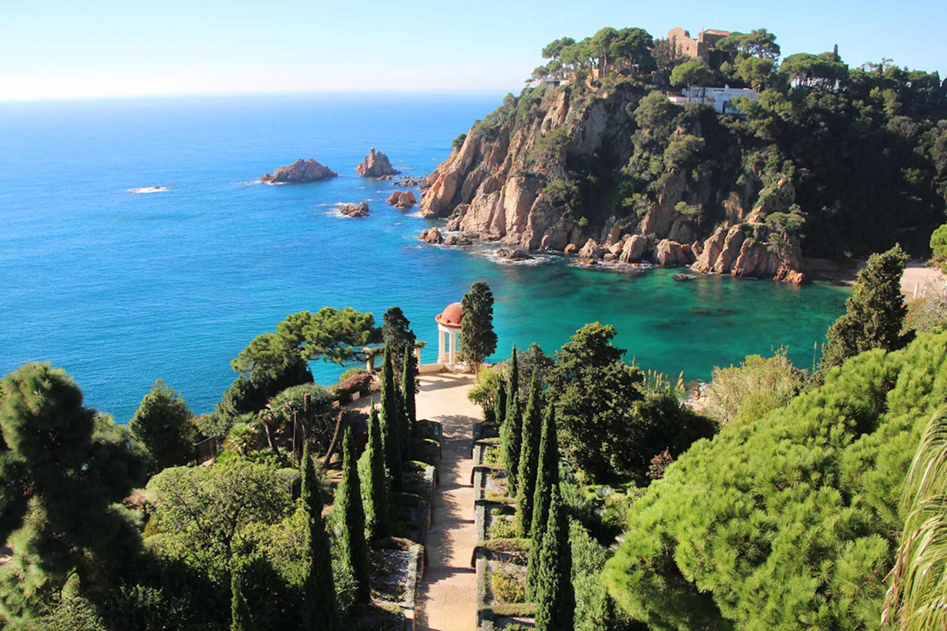 Mediterranean coastal scene with cliffs, turquoise water, and a garden path lined with cypress trees leading to a small gazebo.