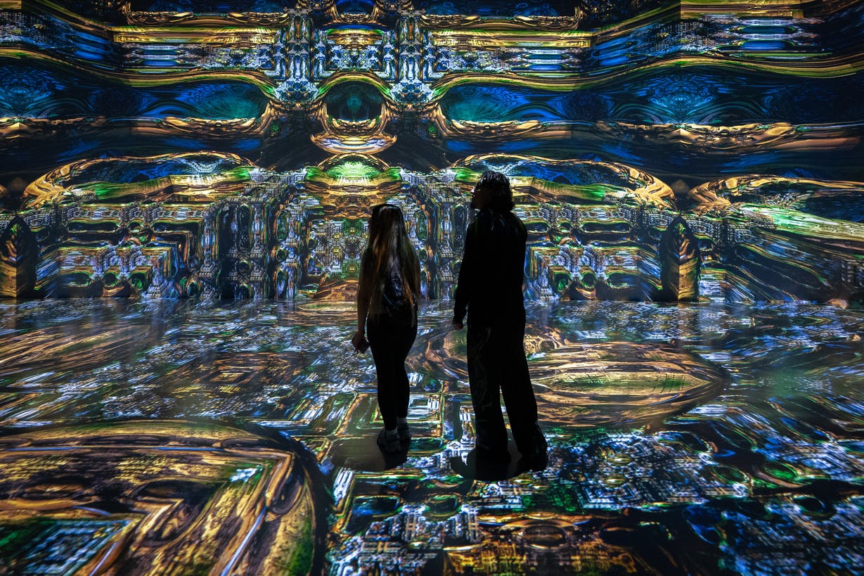 Two people in a colorful digital art installation with vibrant, intricate patterns projected on walls and floor.
