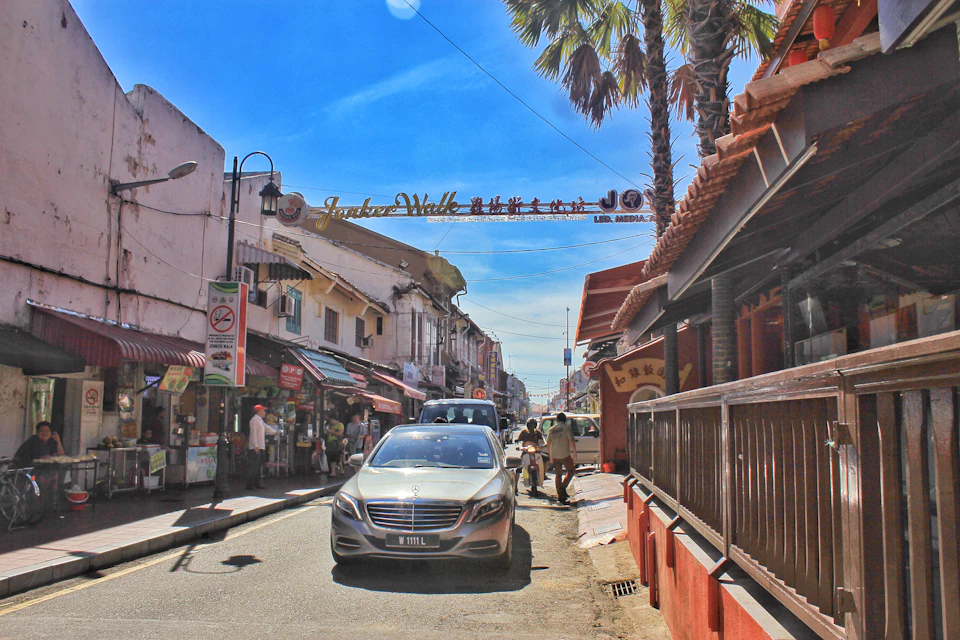 A bustling street with shops on both sides, a car driving under an archway, and a few people walking. Palm trees are visible in the background.