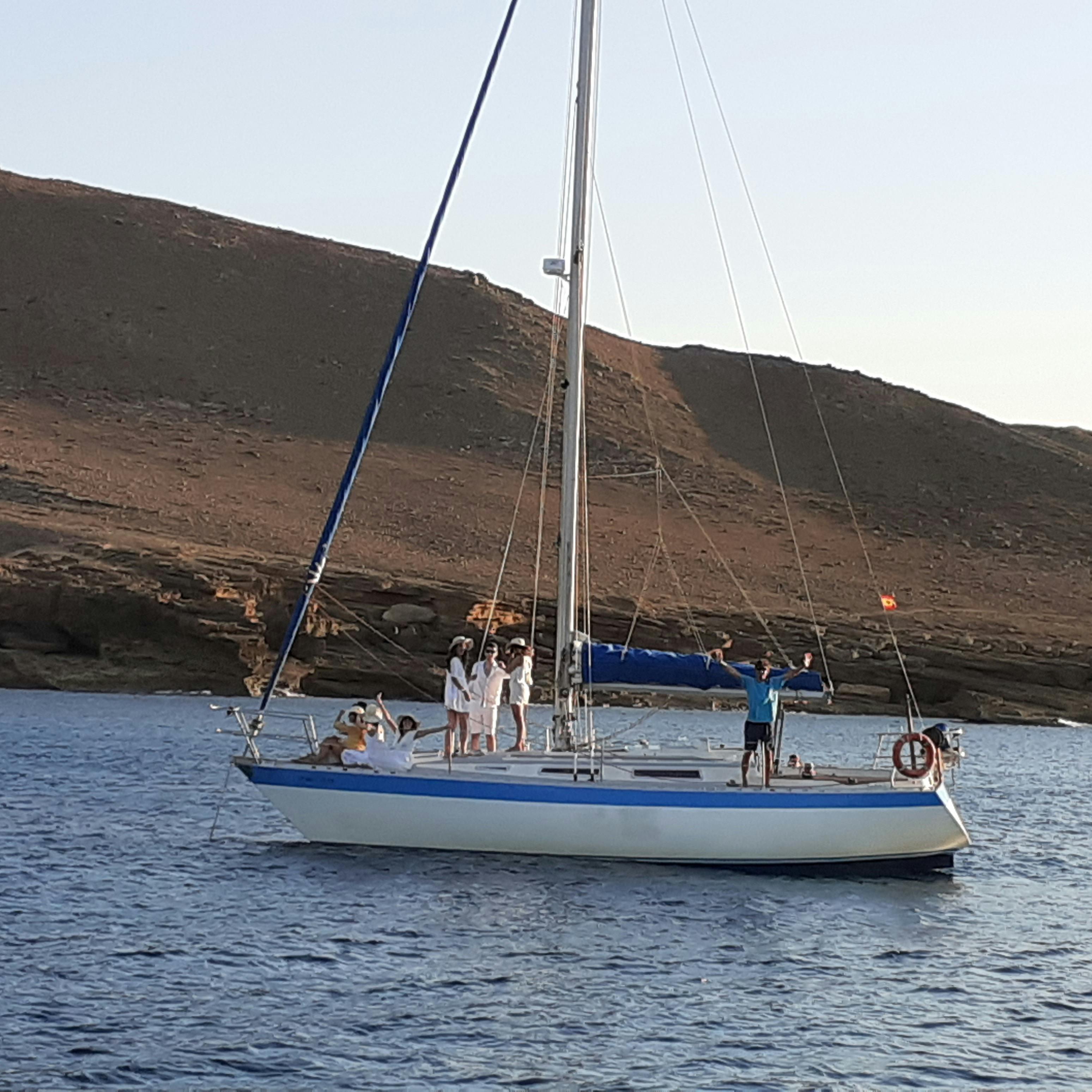 Un groupe de personnes sur un voilier aux voiles blanches et à la couverture bleue, flottant près d'un rivage rocheux sous un ciel clair.
