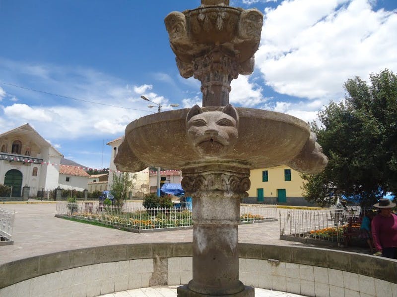 Πέτρινη βρύση με σκαλιστά πρόσωπα ζώων, σε πλακόστρωτη πλατεία με περιμετρικά κτίρια και πράσινο κάτω από έναν εν μέρει νεφελώδη ουρανό.