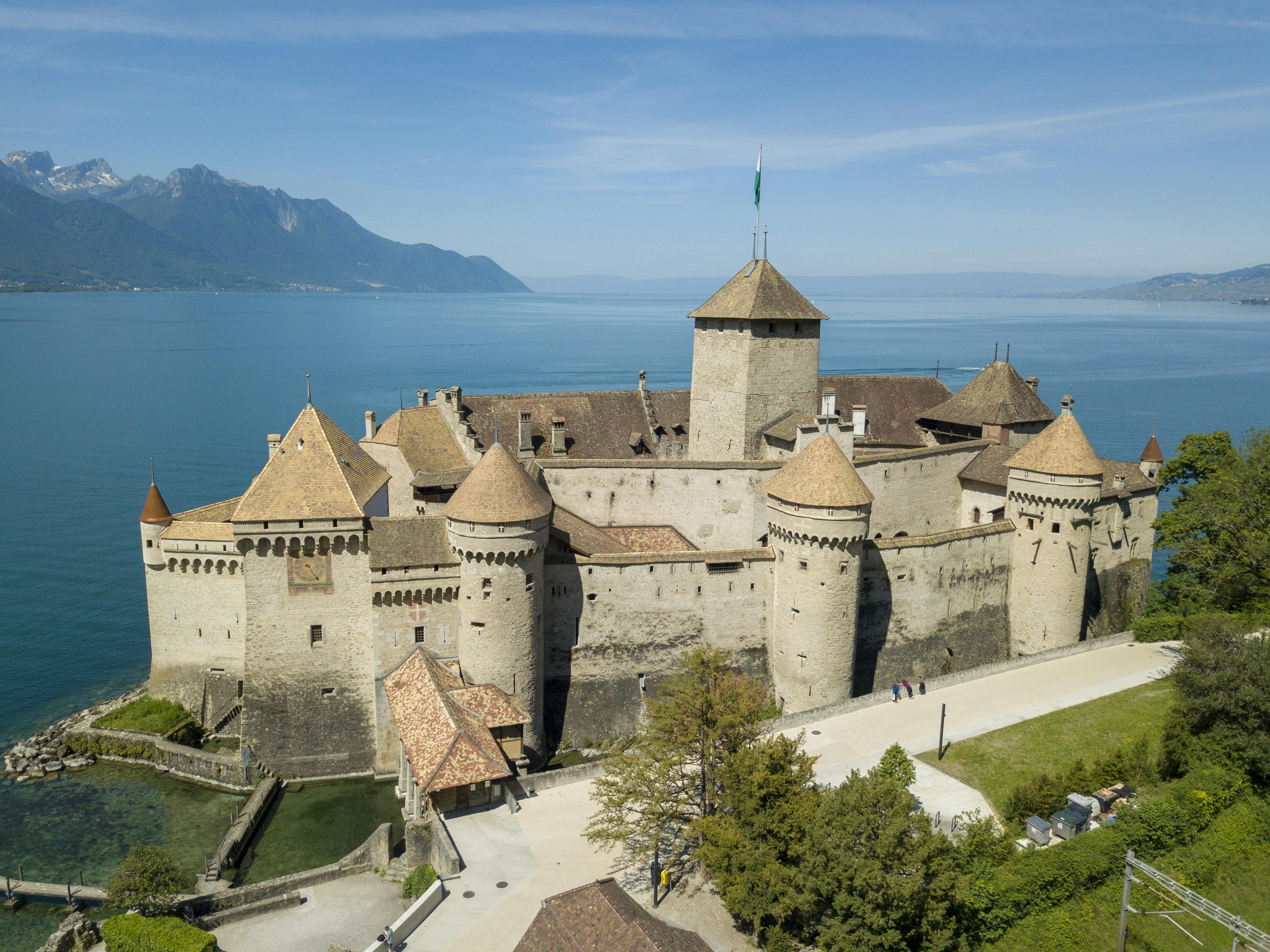 Bird's-eye view of Château de Chillon, a medieval castle on the shore of Lake Geneva, with distant mountains and clear skies.