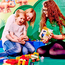 Barn som leker med byggklossar, sitter i en vuxens knä, medan en annan vuxen ler och håller i en bok. Färgglad lekrumsbakgrund.