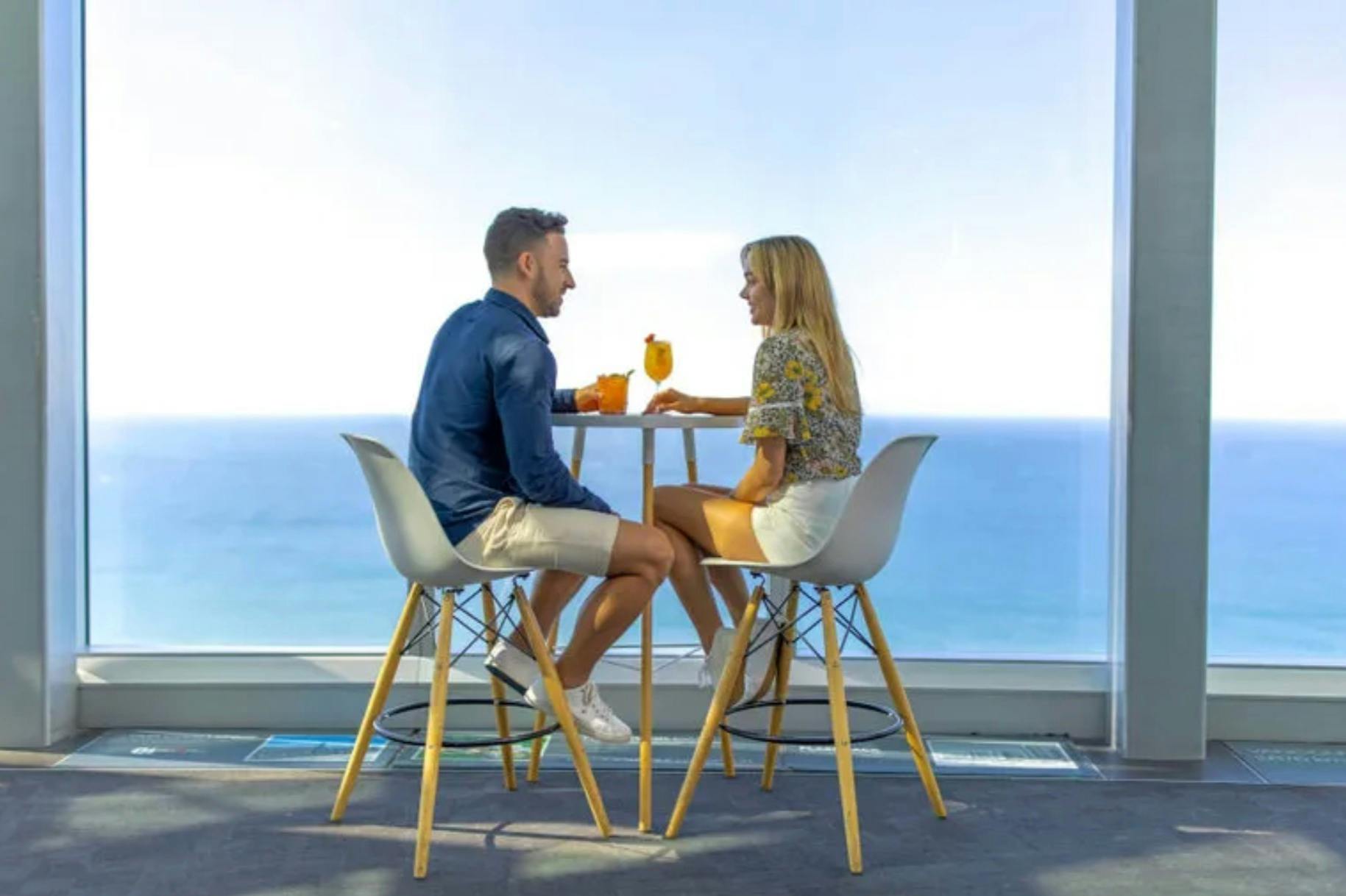 Two people sit at a high table with drinks, overlooking a clear blue ocean through a large window.