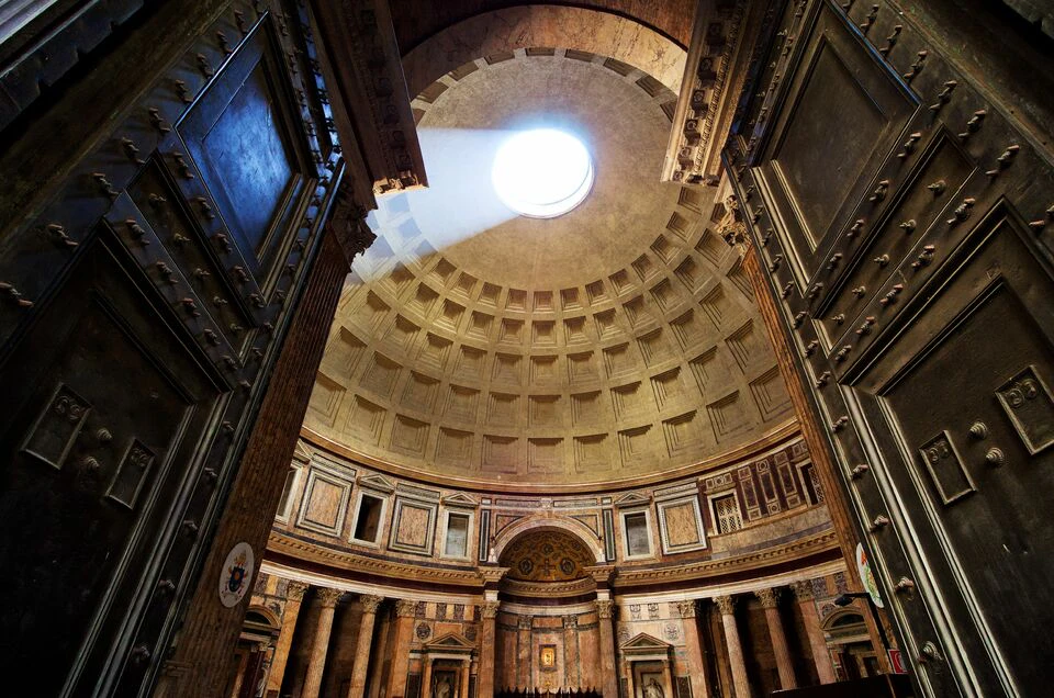 A grand interior with a large domed ceiling and an oculus allowing sunlight to stream in, revealing ornate architectural details.