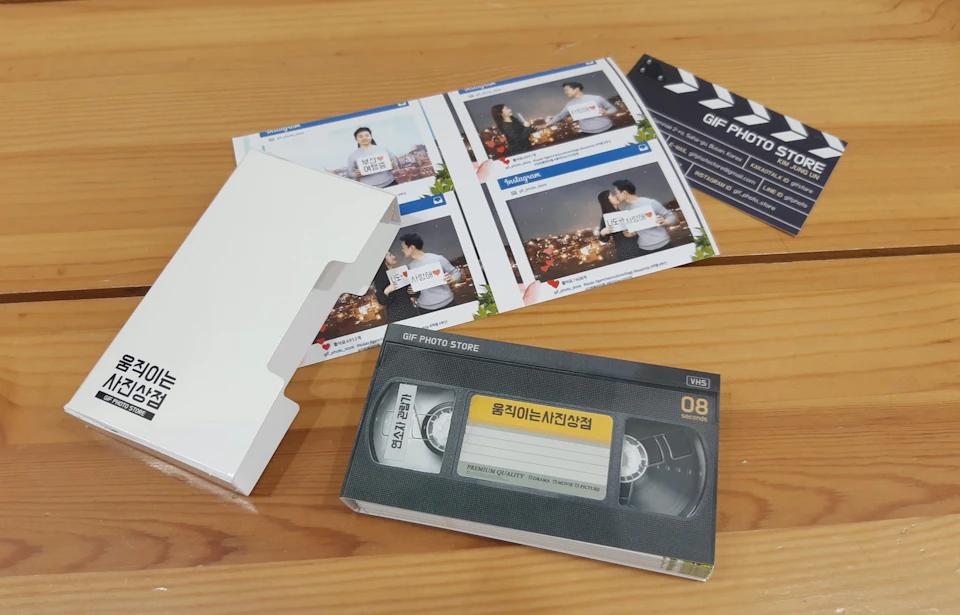 A VHS-style gift box with a label in Korean, printed photo sheets, and a white box on a wooden surface.