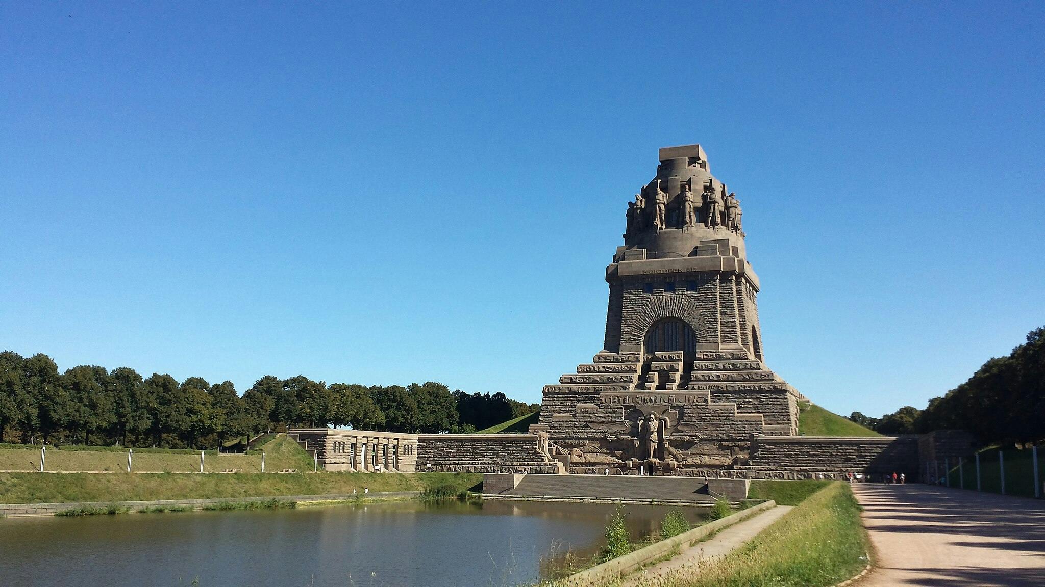 Stone monument with sculptures, surrounded by water and greenery, under a clear blue sky. Few people walking nearby.