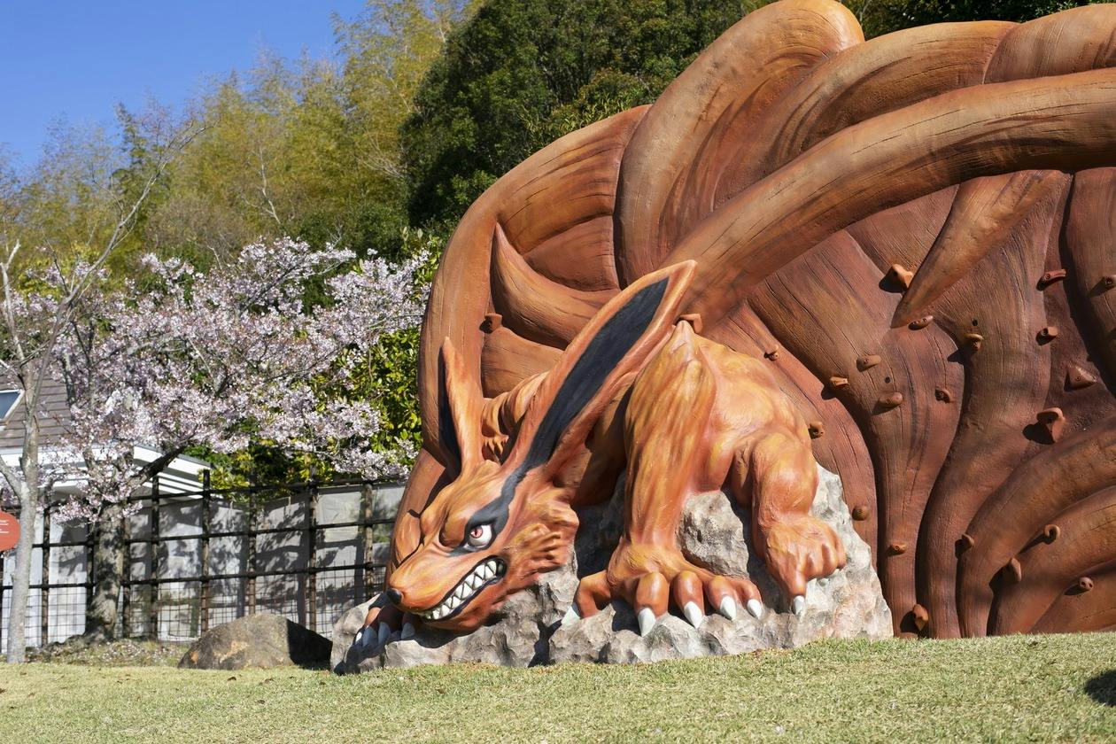 A large, orange fox-like statue with nine tails in a park setting with trees and a wooden fence in the background.