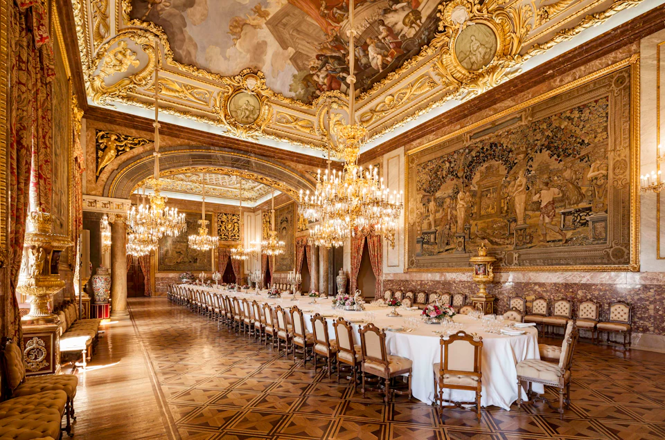 A lavish, ornately decorated dining hall with a long, elegantly set table, multiple chandeliers, and intricate wall and ceiling art.