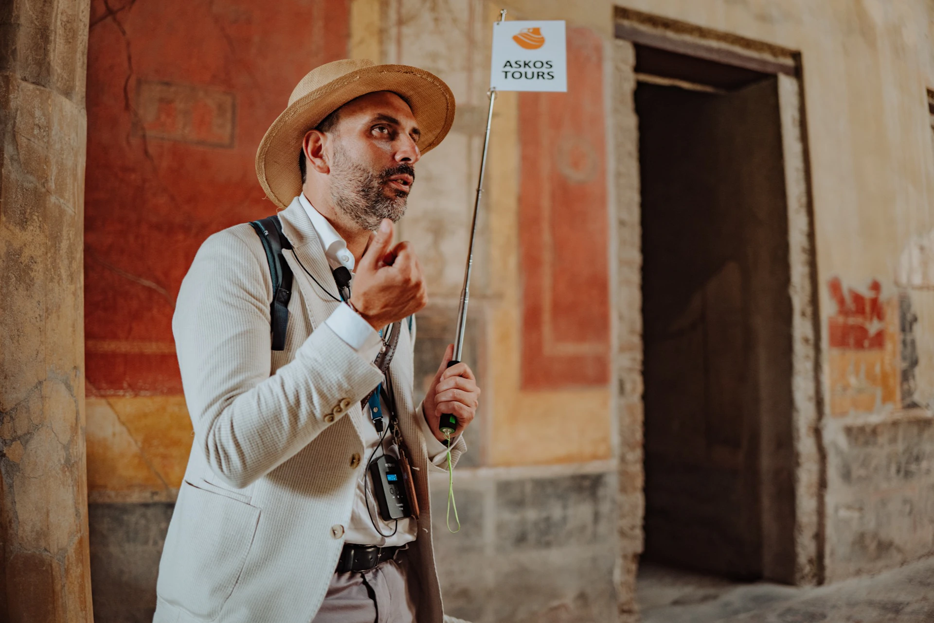 A tour guide wearing a hat and holding a sign that reads "Askos Tours" stands in front of a painted wall indoors.