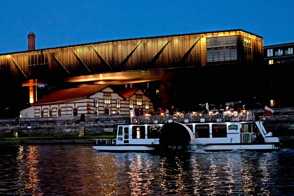 A white riverboat travels past a lit, modern bridge and a historic building at dusk. Reflections shimmer on the water.