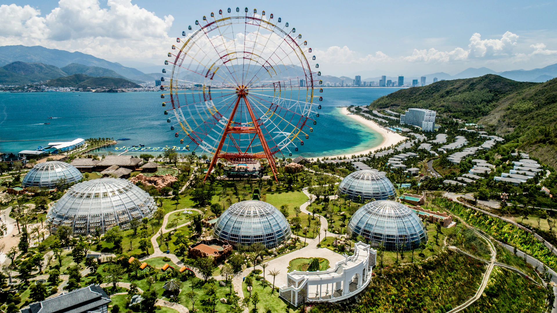 A large ferris wheel near a beach, surrounded by domed structures and landscaped greenery, with a cityscape in the background.