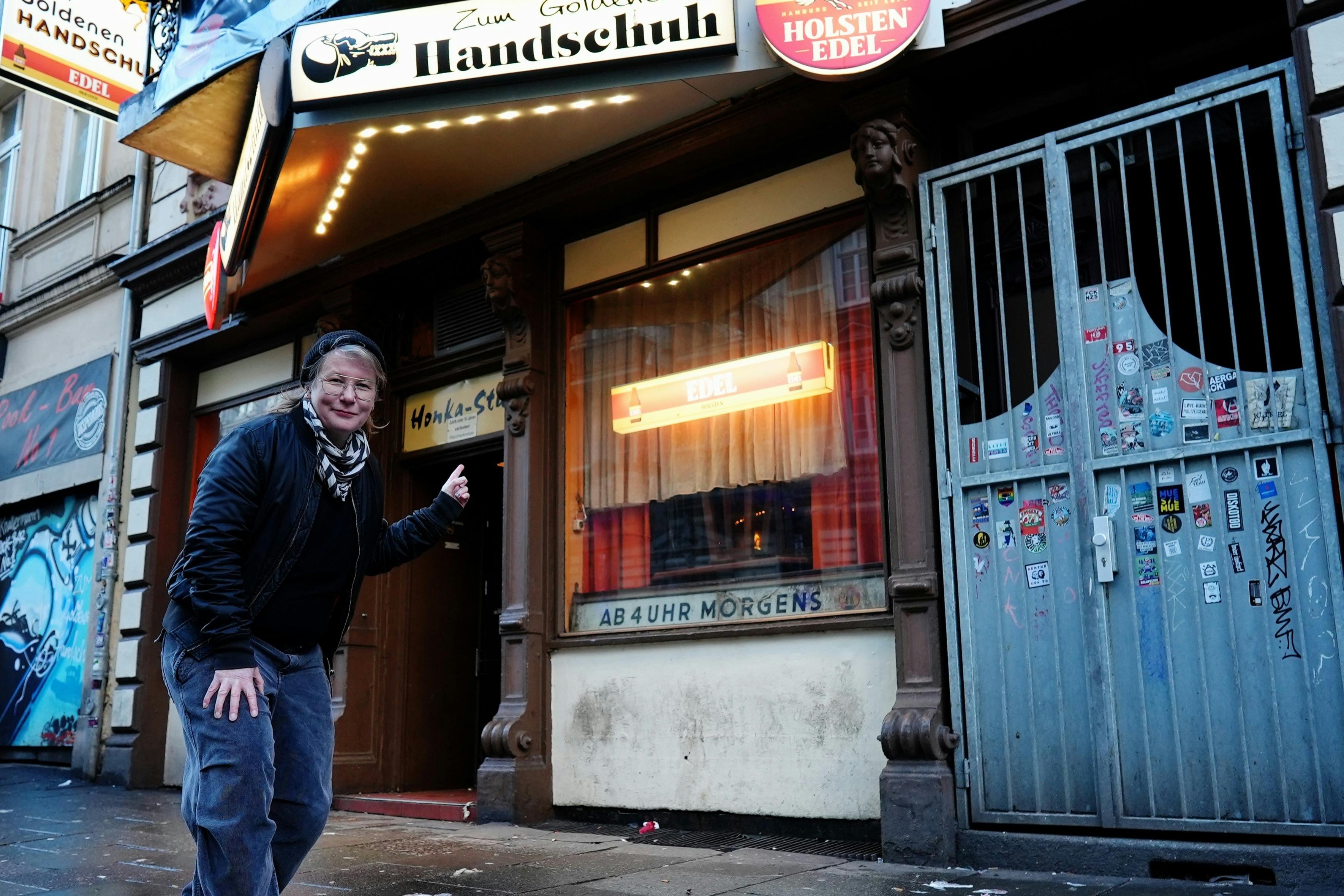 En person står uden for en gadevendt forretning med skiltning med teksten "Zum Goldenen Handschuh." De peger mod indgangen.