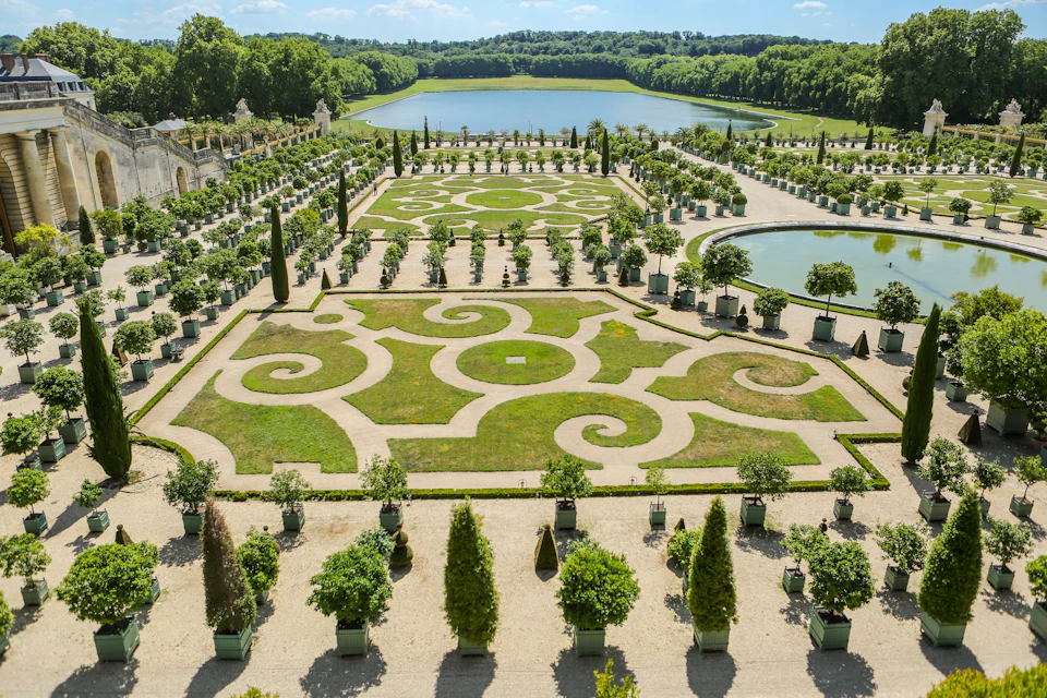 A formal garden with symmetrical patterns, potted trees, and a large water feature in the background.
