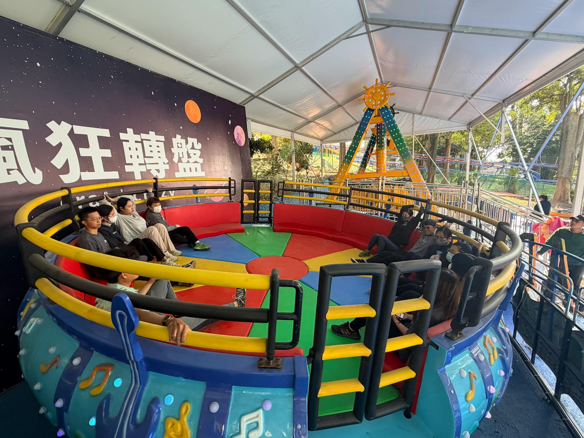People seated on a colorful spinning amusement ride under a tent, with a space-themed backdrop and Chinese text.