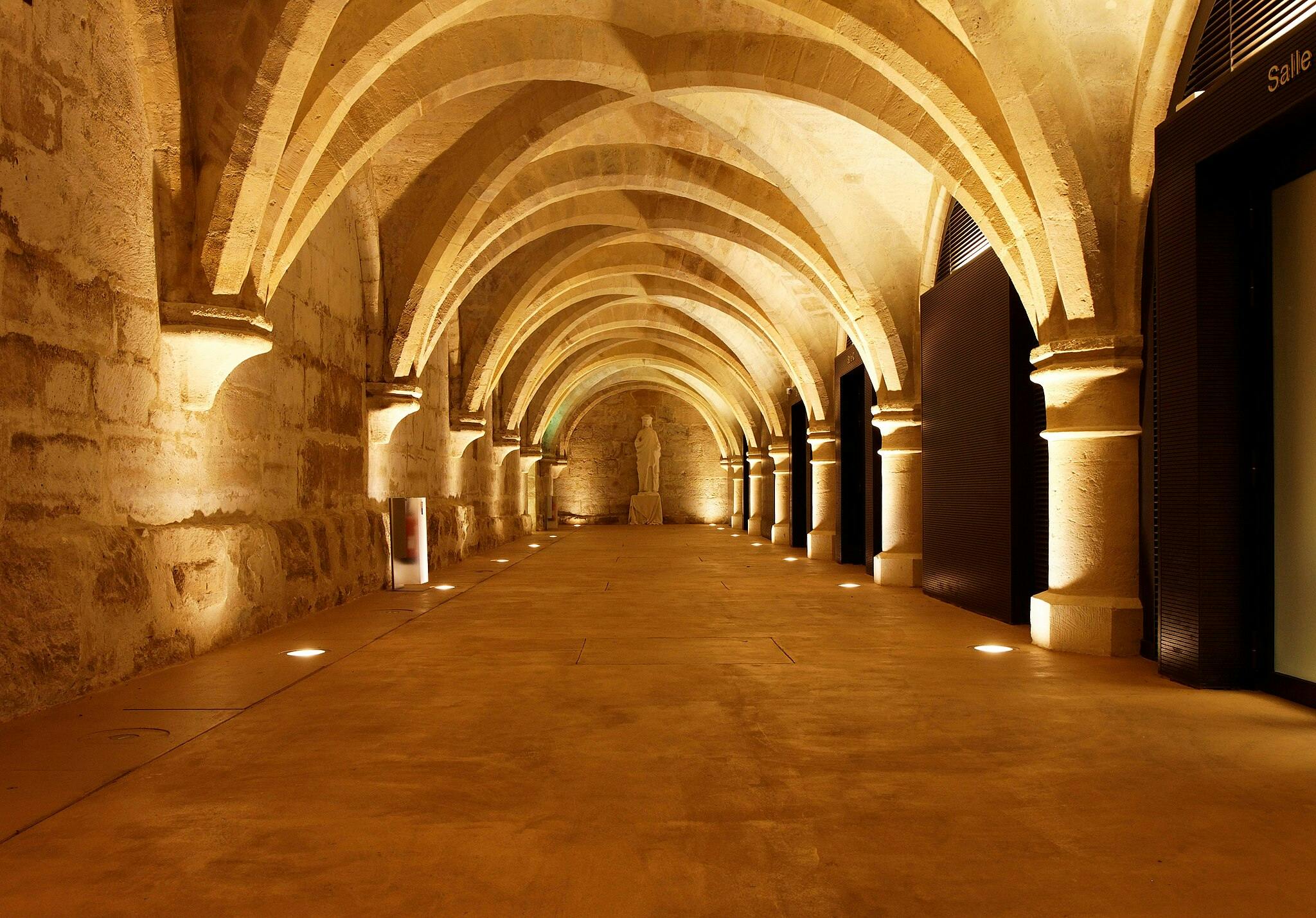 오른쪽에 기둥과 문이 늘어선 아치형 아치형 통로가 있는 아치형 아치형 석조 복도로, 끝에는 흰색 동상이 있습니다.