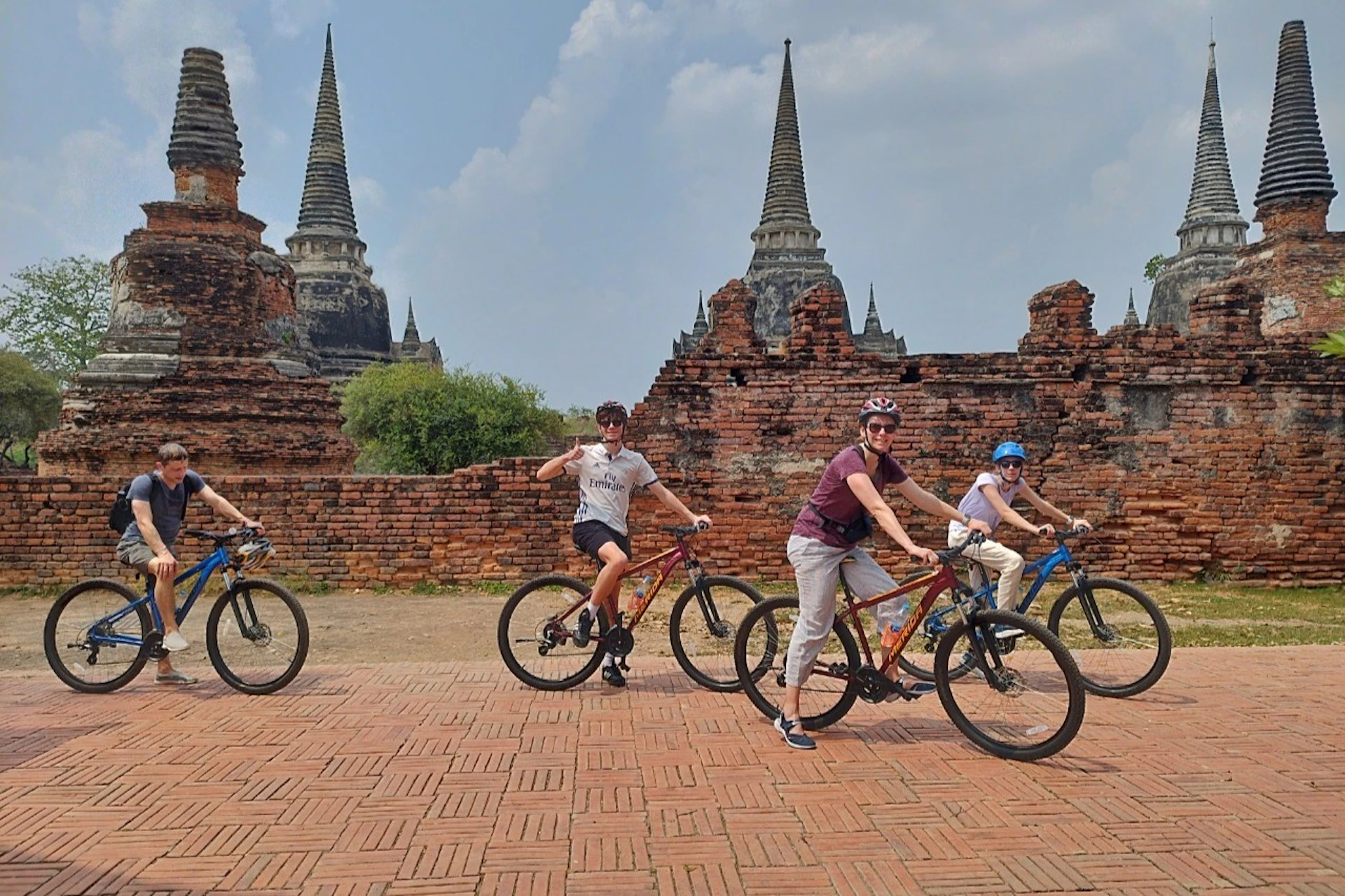 Four people riding bicycles in a historical site with ancient brick walls and spired structures in the background.