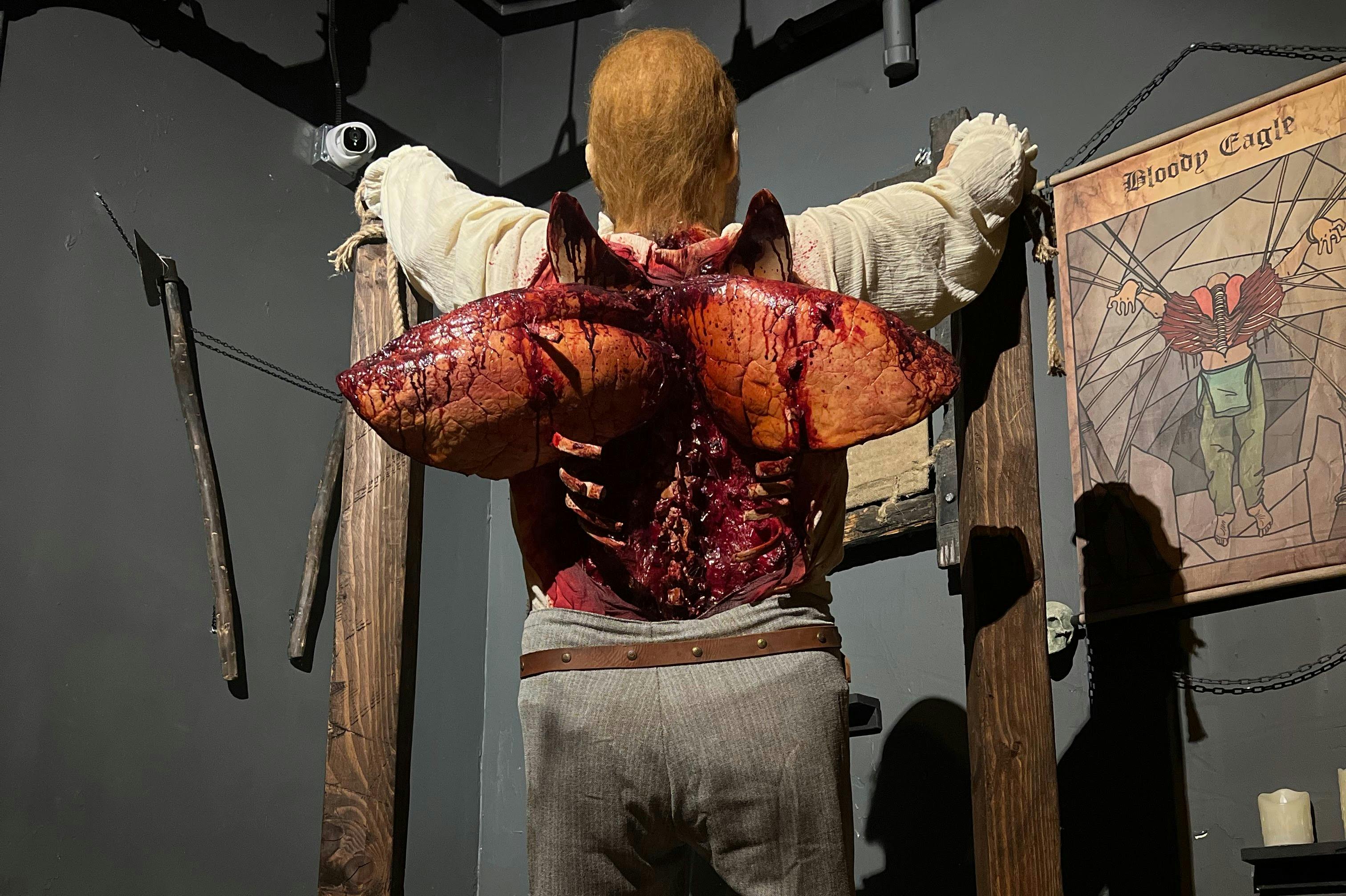 Uma figura com grandes e grotescas asas a sair das costas, numa sala pouco iluminada com molduras de madeira.