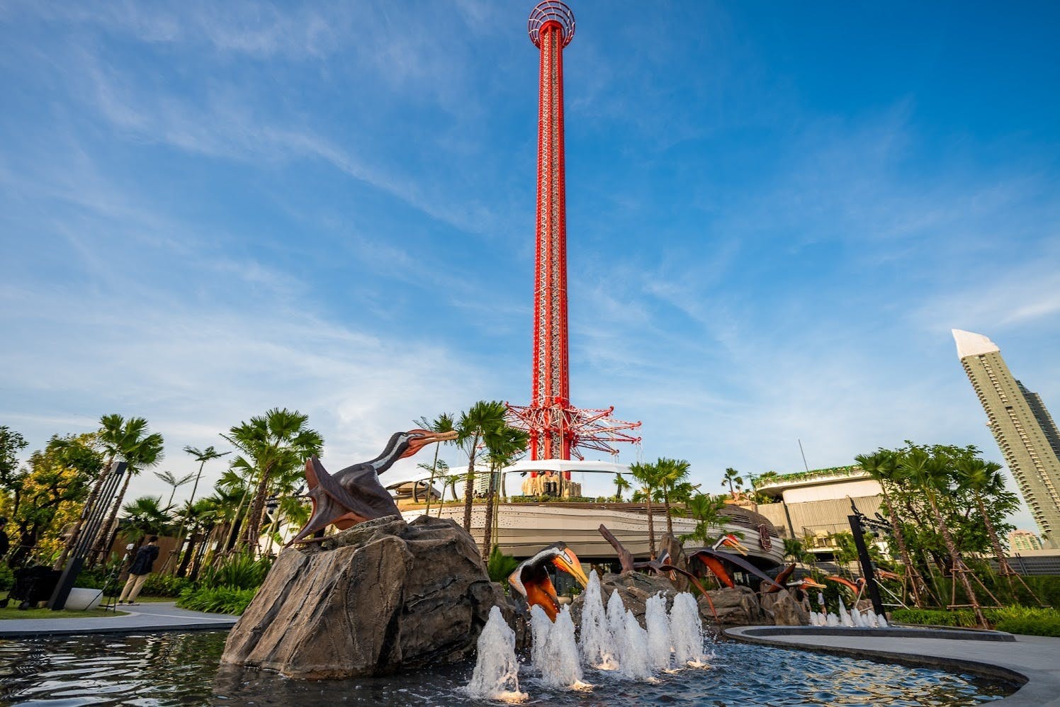 A tall red amusement ride tower surrounded by fountain features, rock sculptures, and palm trees under a clear blue sky.