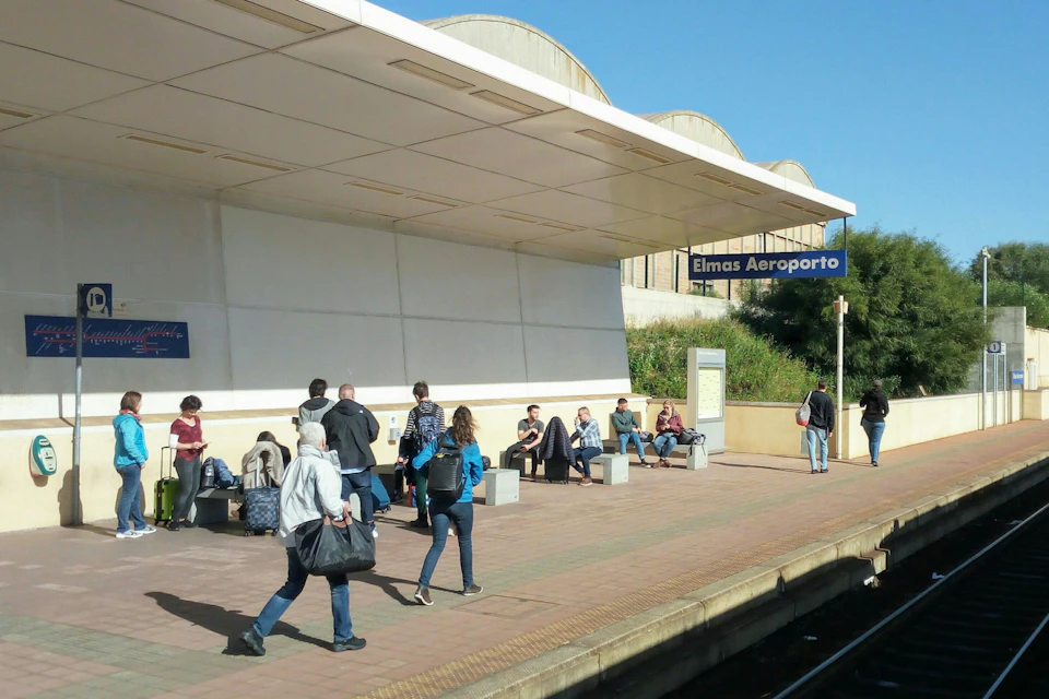 People wait and walk on a platform at Elmas Aeroporto train station under a clear sky, with benches and signage visible.
