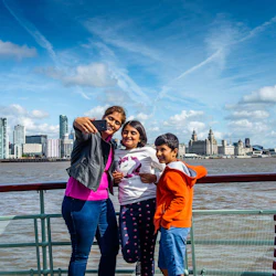 Tre personer ler och tar en selfie på en båt med en stads skyskrapor och vattnet bakom sig, under en klarblå himmel.