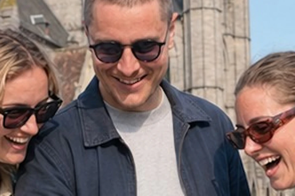 Three people smiling while looking at a phone, standing in front of a Gothic-style cathedral on a sunny day.