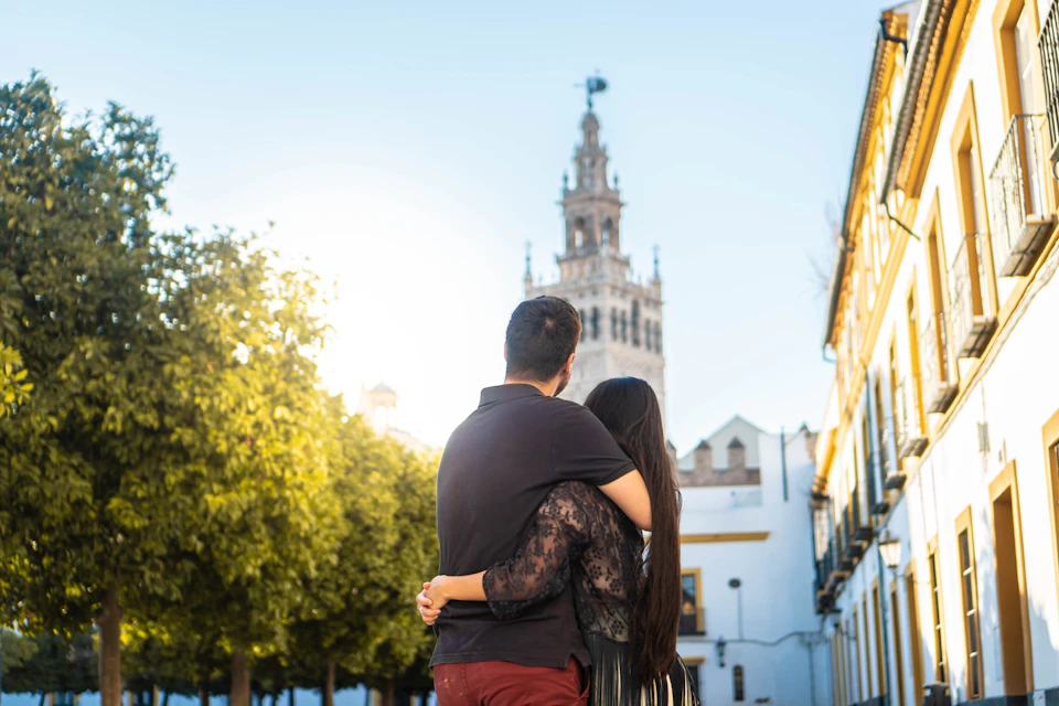 A couple embraces while looking at a tall, ornate tower in a sunny, tree-lined urban setting.
