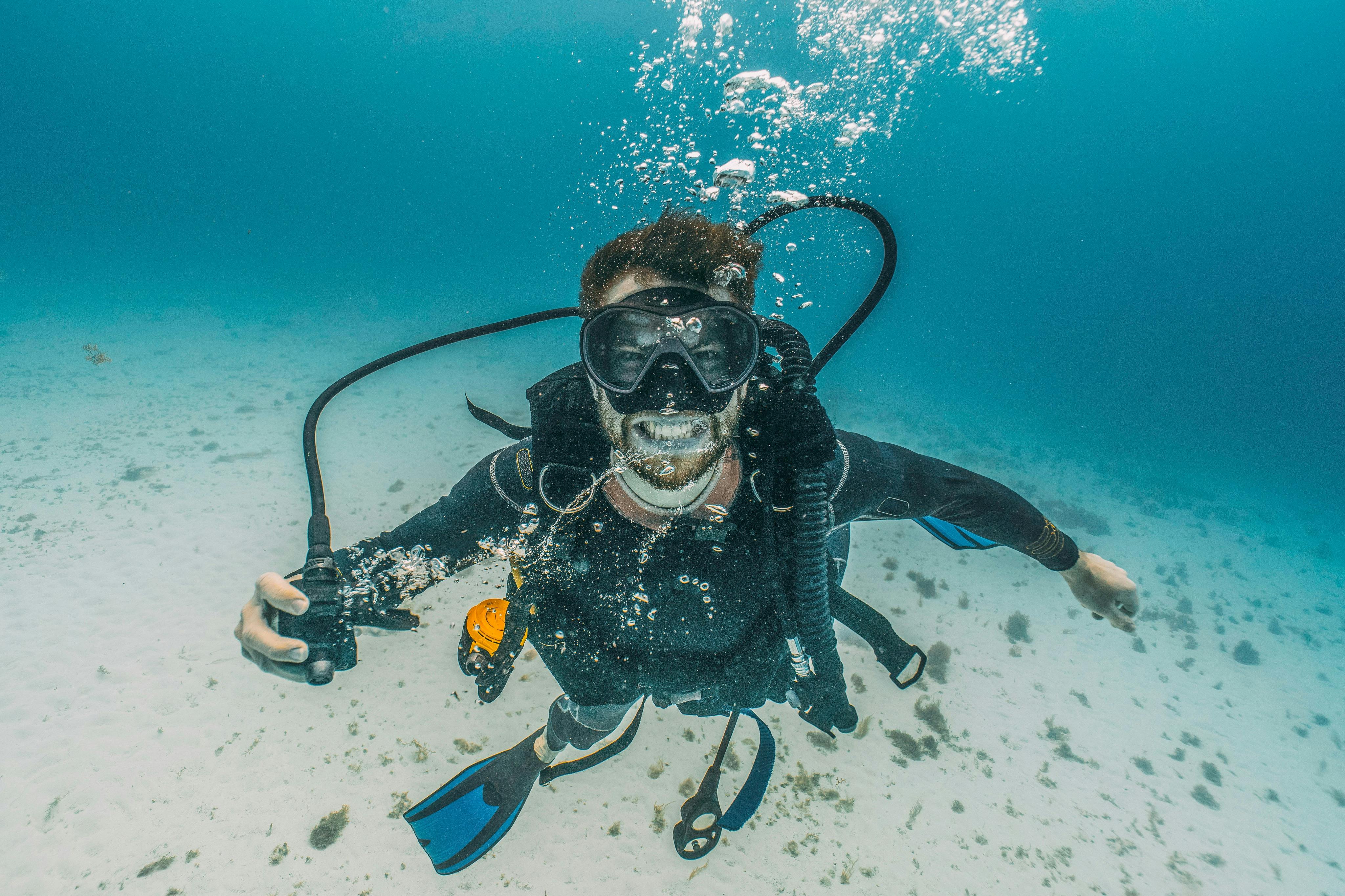 Plongeur sous-marin, vêtu d'une combinaison, d'un masque et de palmes, avec des bulles d'air autour de lui. Le fond de l'océan est visible en dessous.
