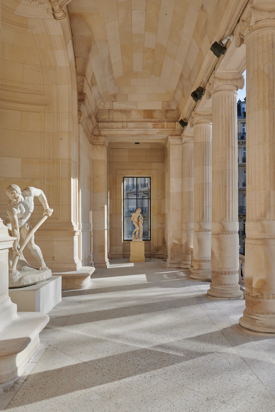 A sunlit colonnade with classical statues, including a man bending with a tool and a distant standing figure.