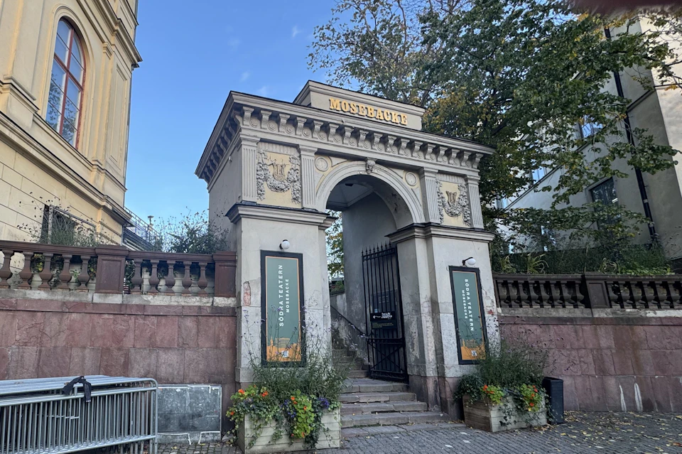 An ornate stone gateway labeled "Monumentet" with stairs leading up, flanked by planters and decorated with carvings.