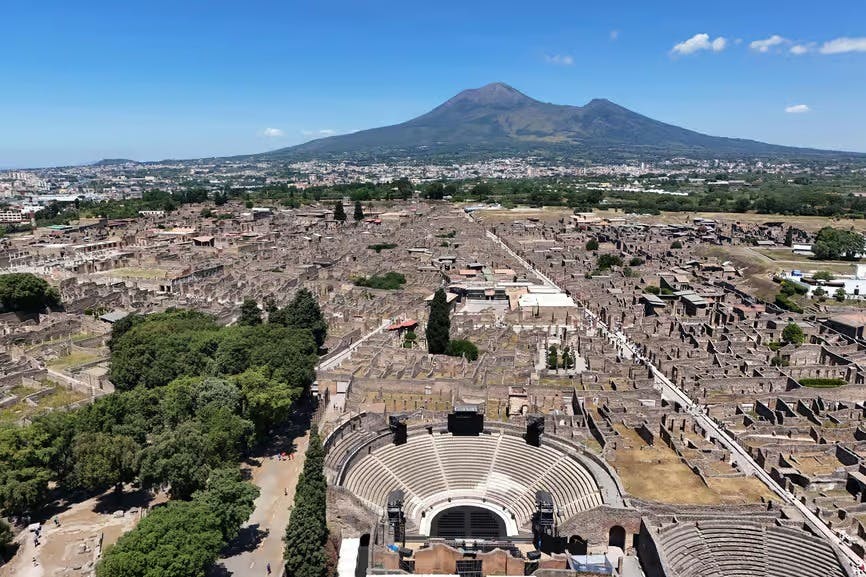 鸟瞰古代遗址，包括一个圆形剧场，周围环绕着绿色的树木，背景是一座晴朗的天空下的大山。