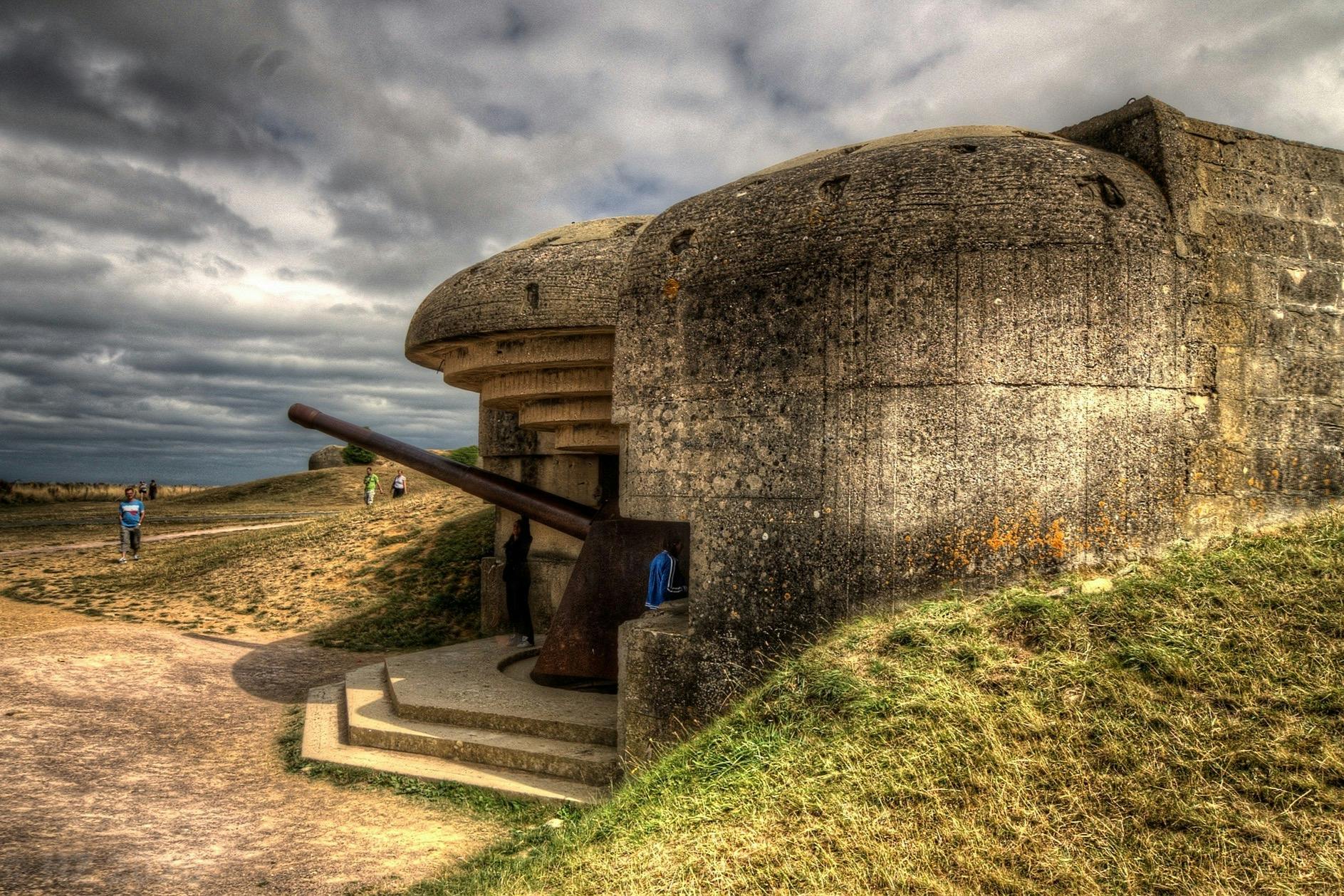 A historic concrete bunker with a large cannon, surrounded by grass and pathways under a cloudy sky.